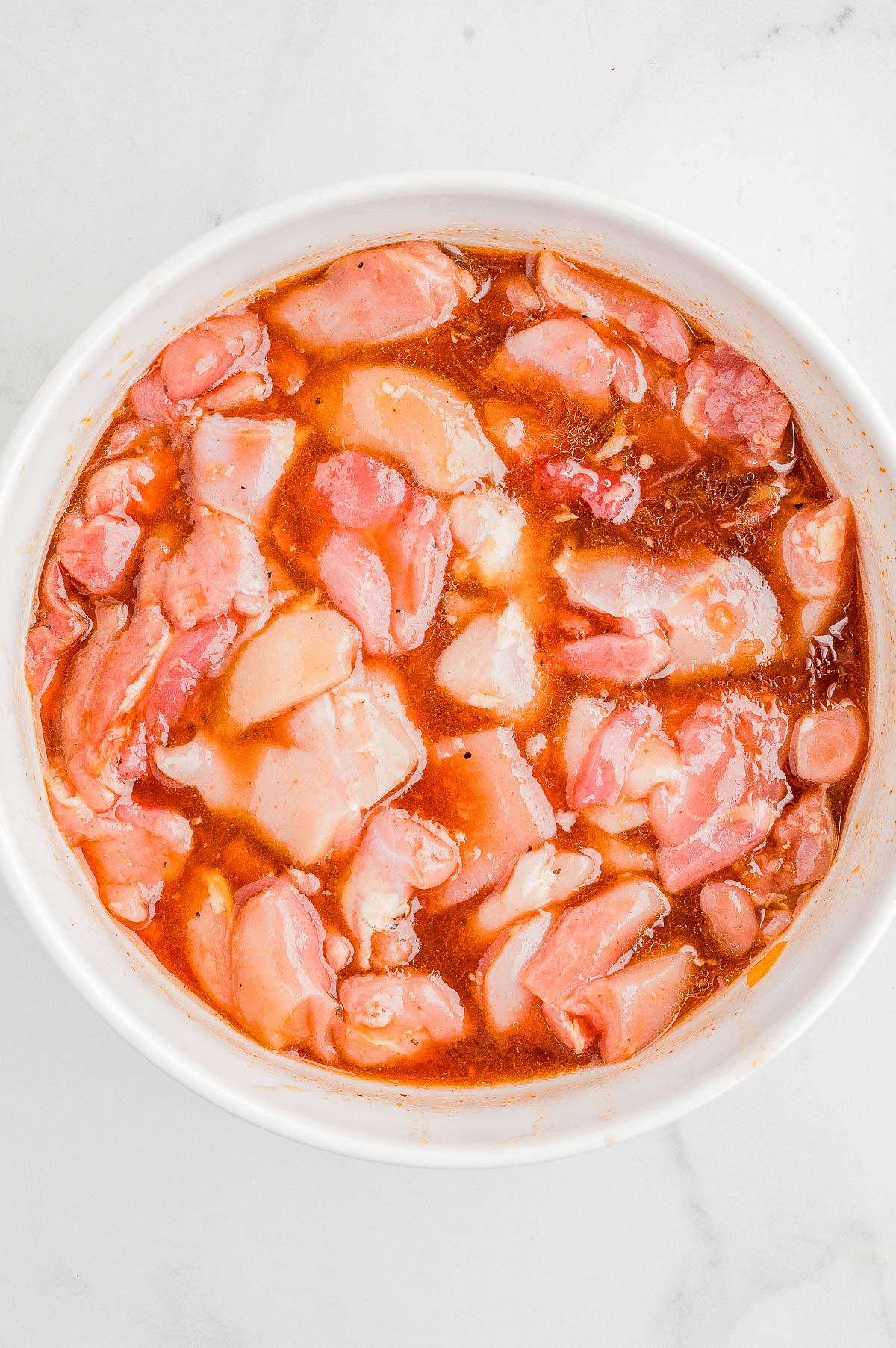 Raw chicken pieces marinating in a reddish-brown liquid inside a white bowl on a white surface.
