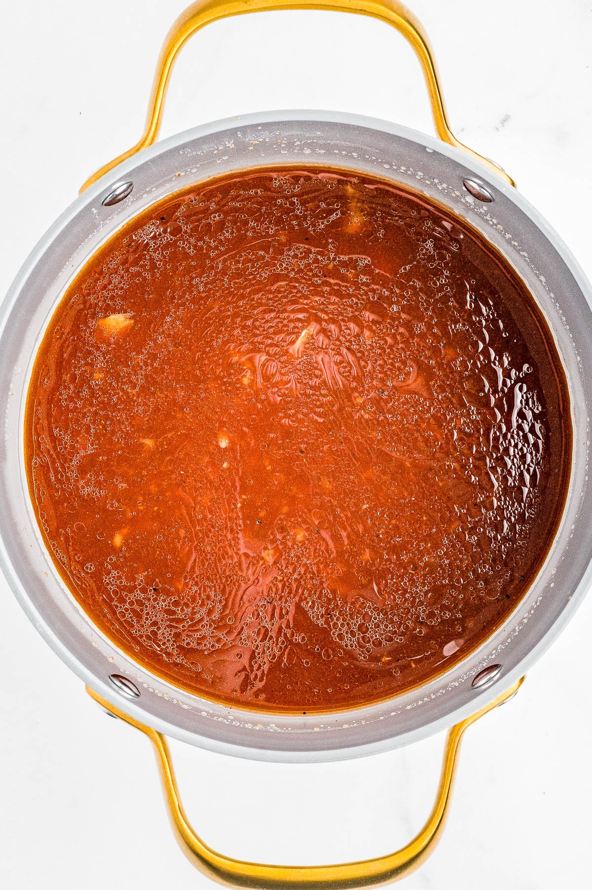 A pot filled with simmering red tomato-based sauce, viewed from above, with gold handles on either side.