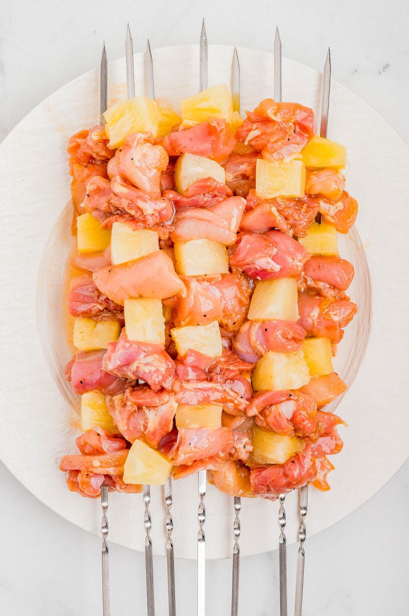 Five metal skewers with alternating pieces of raw marinated chicken and pineapple chunks arranged on a white plate.