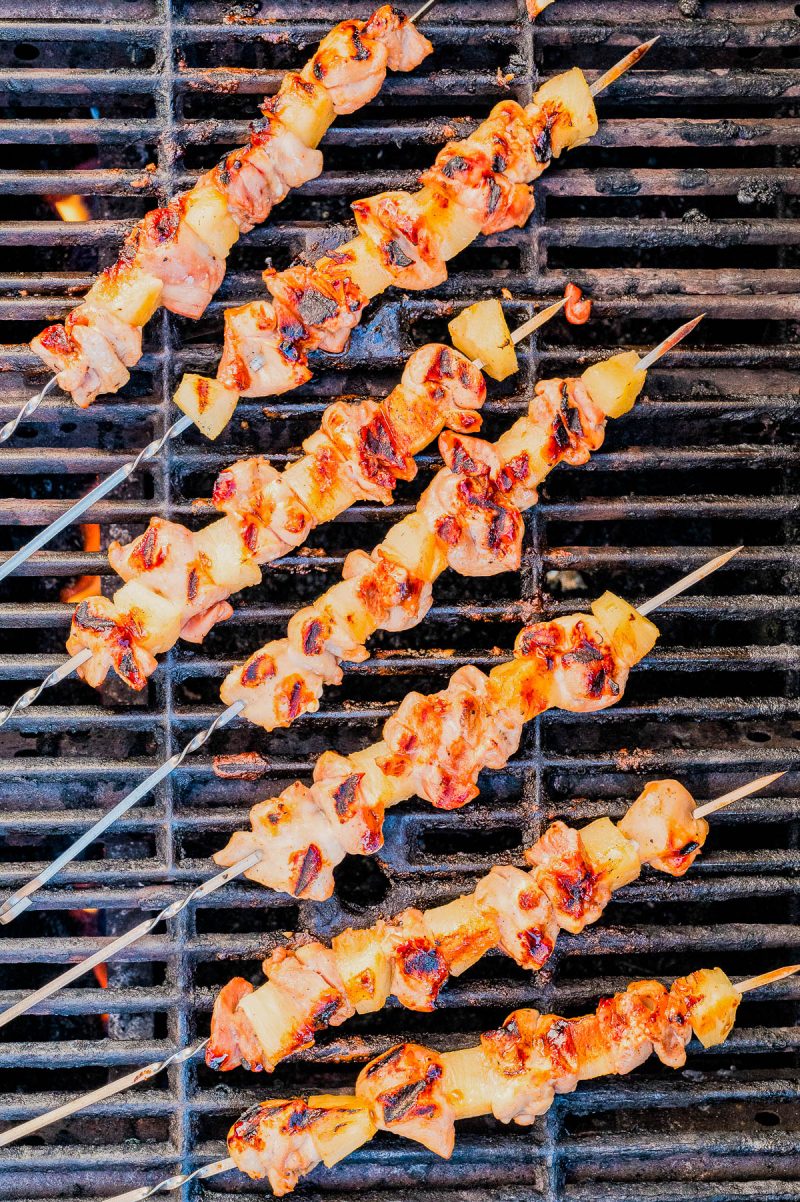 Seven skewers of grilled meat and pineapple are cooking on a metal barbecue grill, with some pieces showing char marks.