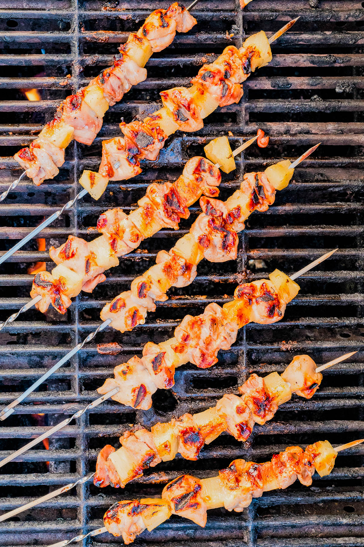 Seven skewers of grilled meat and pineapple are cooking on a metal barbecue grill, with some pieces showing char marks.