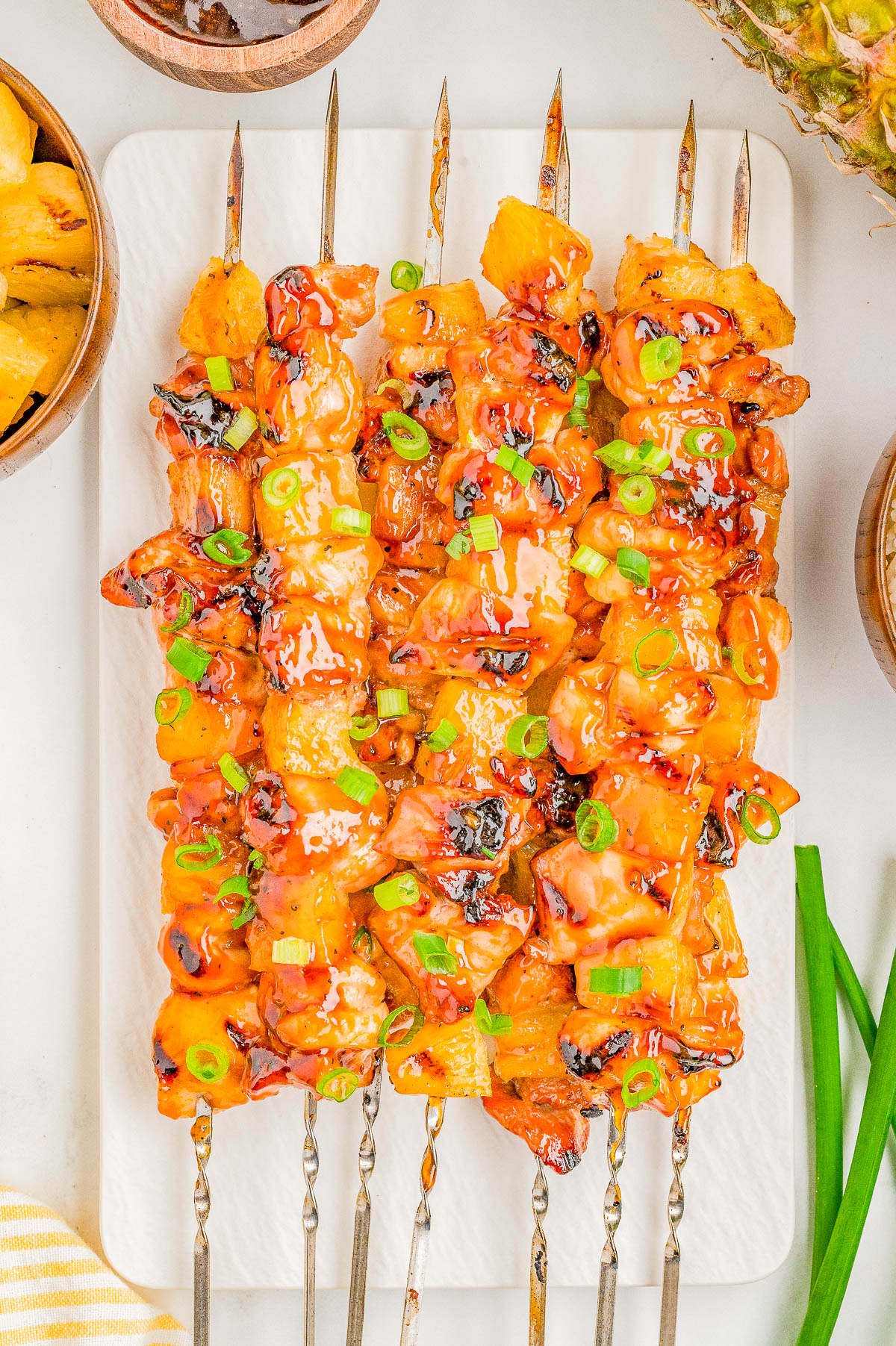 Six metal skewers with grilled chicken and pineapple pieces, glazed with sauce and garnished with chopped green onions, arranged on a white serving board.