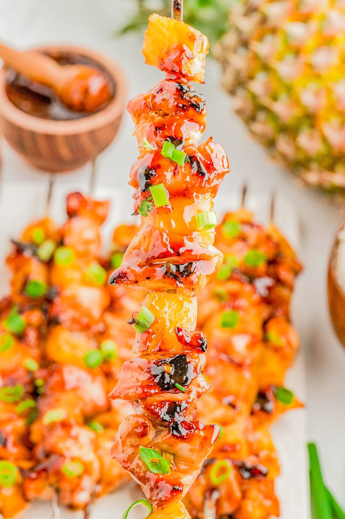 A close-up of a grilled chicken and pineapple skewer topped with chopped green onions and glaze, with more skewers and a pineapple in the background.
