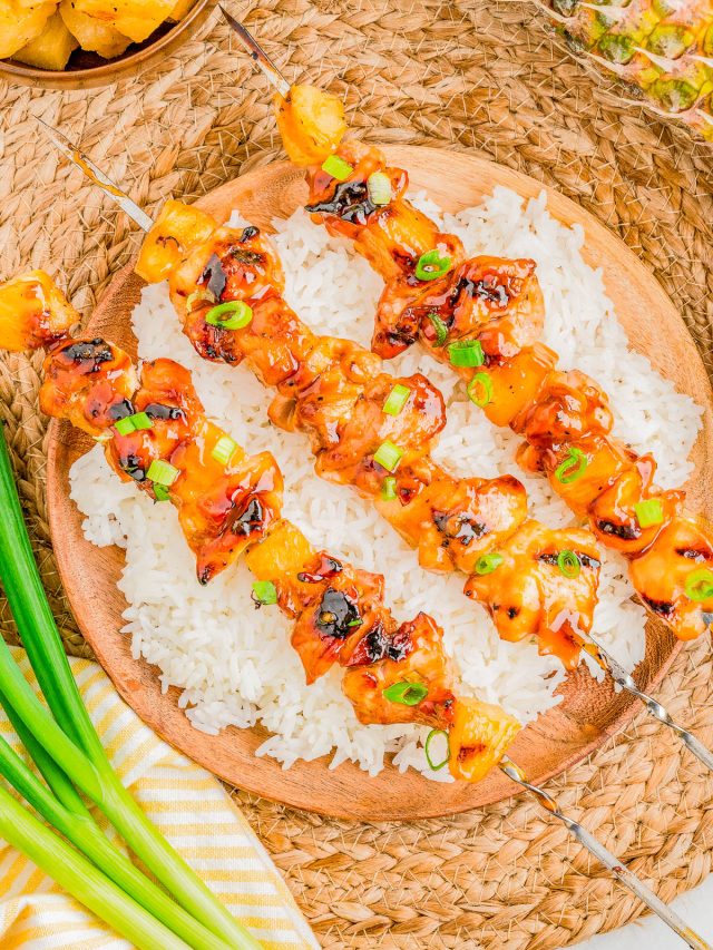 Three grilled chicken and pineapple skewers are served on a bed of white rice, garnished with chopped green onions, with a side of green onions and pineapple pieces nearby.