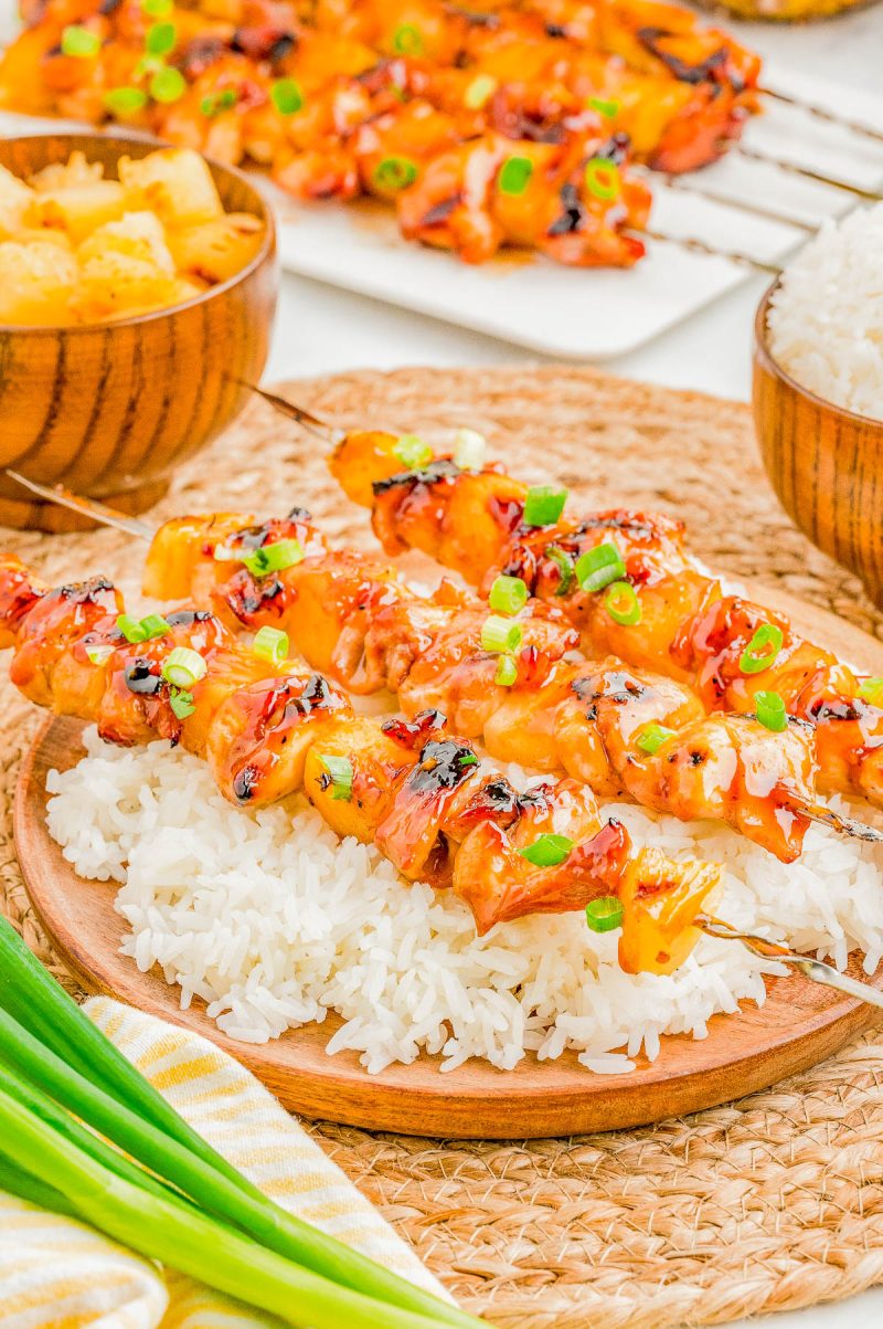 Grilled chicken and pineapple skewers garnished with green onions, served on a bed of white rice with bowls of rice and pineapple in the background.