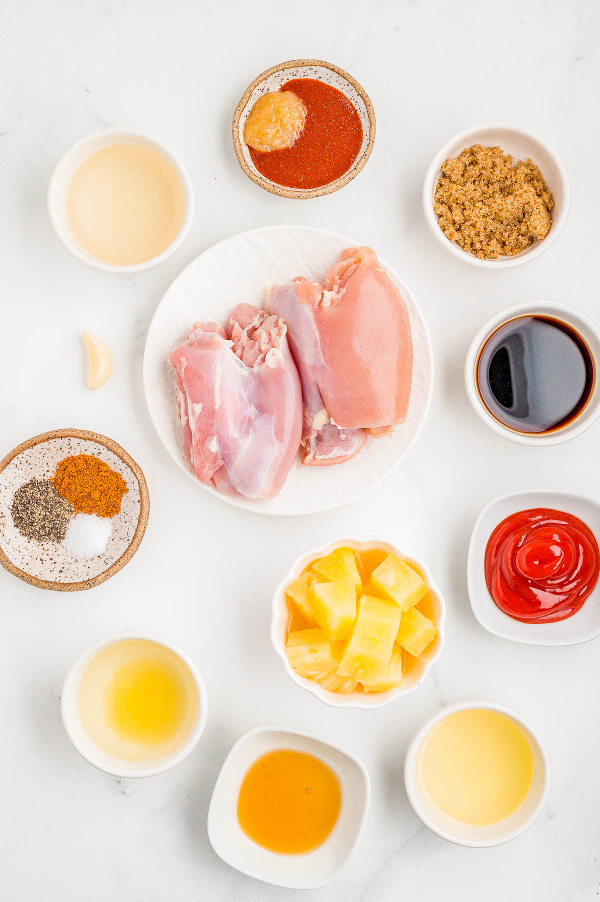 Raw chicken breasts on a plate surrounded by small bowls containing spices, sauces, brown sugar, chopped pineapple, vinegar, and minced garlic on a white surface.