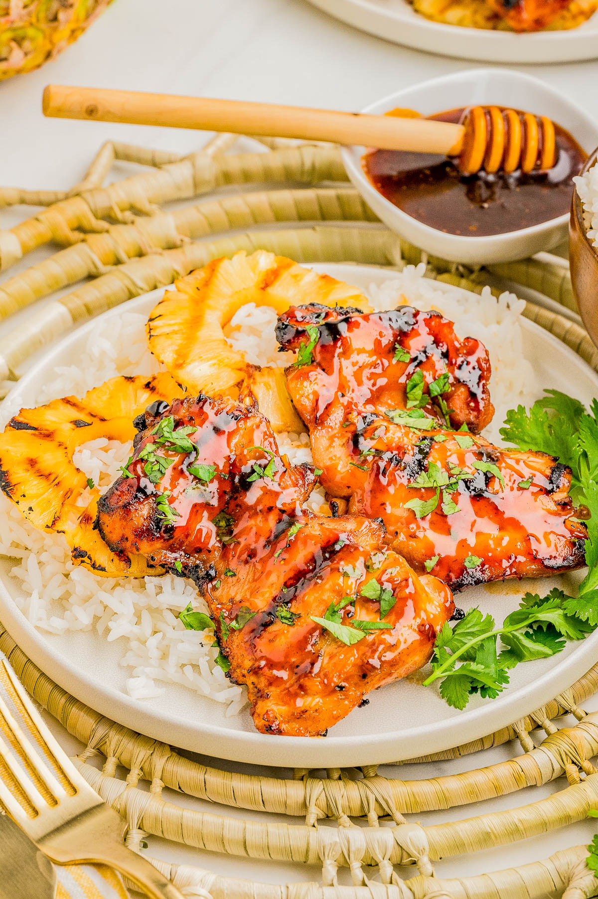 Grilled chicken with glaze served on white rice, garnished with cilantro and grilled pineapple slices, with a small bowl of sauce and honey dipper in the background.