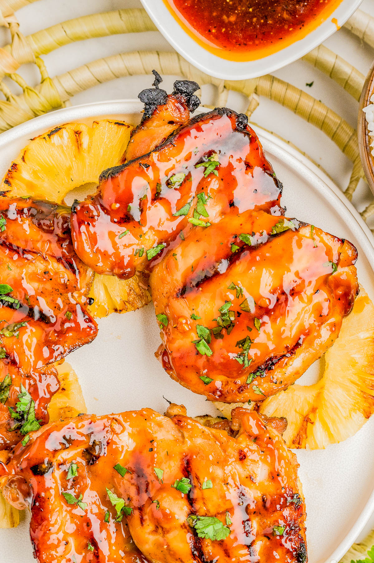 Grilled chicken thighs glazed with sauce, garnished with chopped herbs, served on pineapple slices on a white plate; a bowl of sauce is visible in the background.