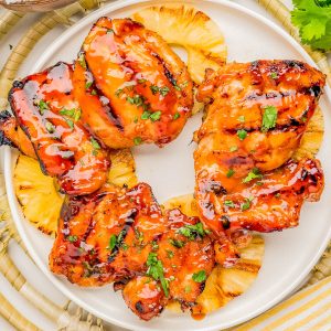 Grilled glazed chicken thighs served on top of pineapple slices, garnished with chopped herbs, on a white plate with rice and fresh cilantro nearby.