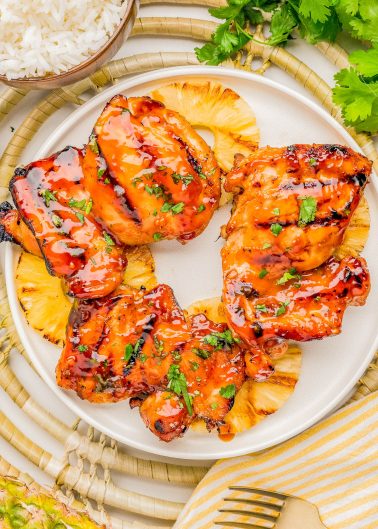 Grilled glazed chicken thighs served on top of pineapple slices, garnished with chopped herbs, on a white plate with rice and fresh cilantro nearby.