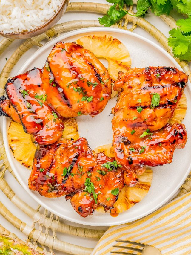 Grilled glazed chicken thighs served on top of pineapple slices, garnished with chopped herbs, on a white plate with rice and fresh cilantro nearby.