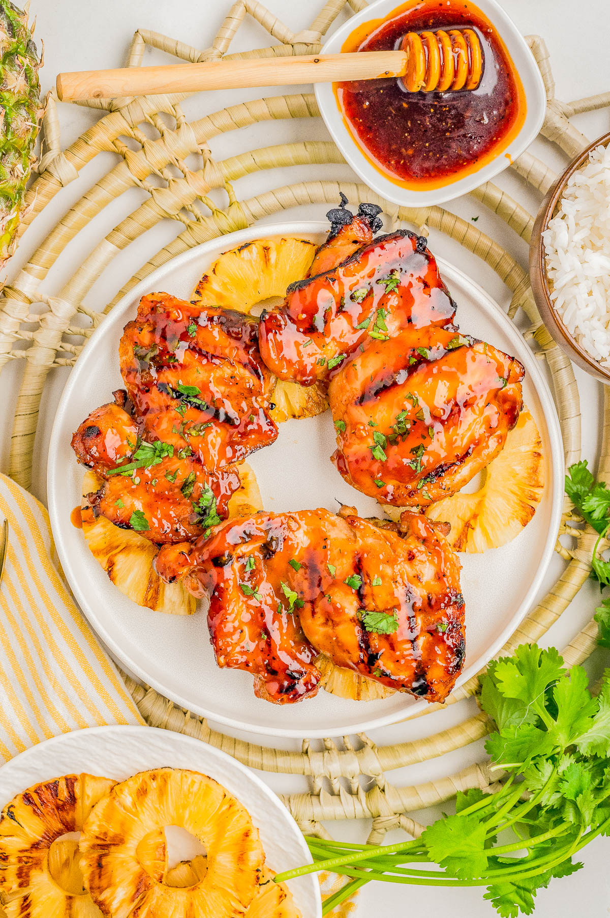 Grilled chicken thighs glazed with sauce, served on pineapple slices, garnished with herbs. A honey dipper, sauce, rice, and fresh cilantro are arranged nearby.