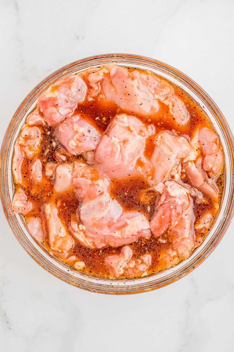 Raw chicken pieces marinating in a bowl with a reddish marinade on a white surface.