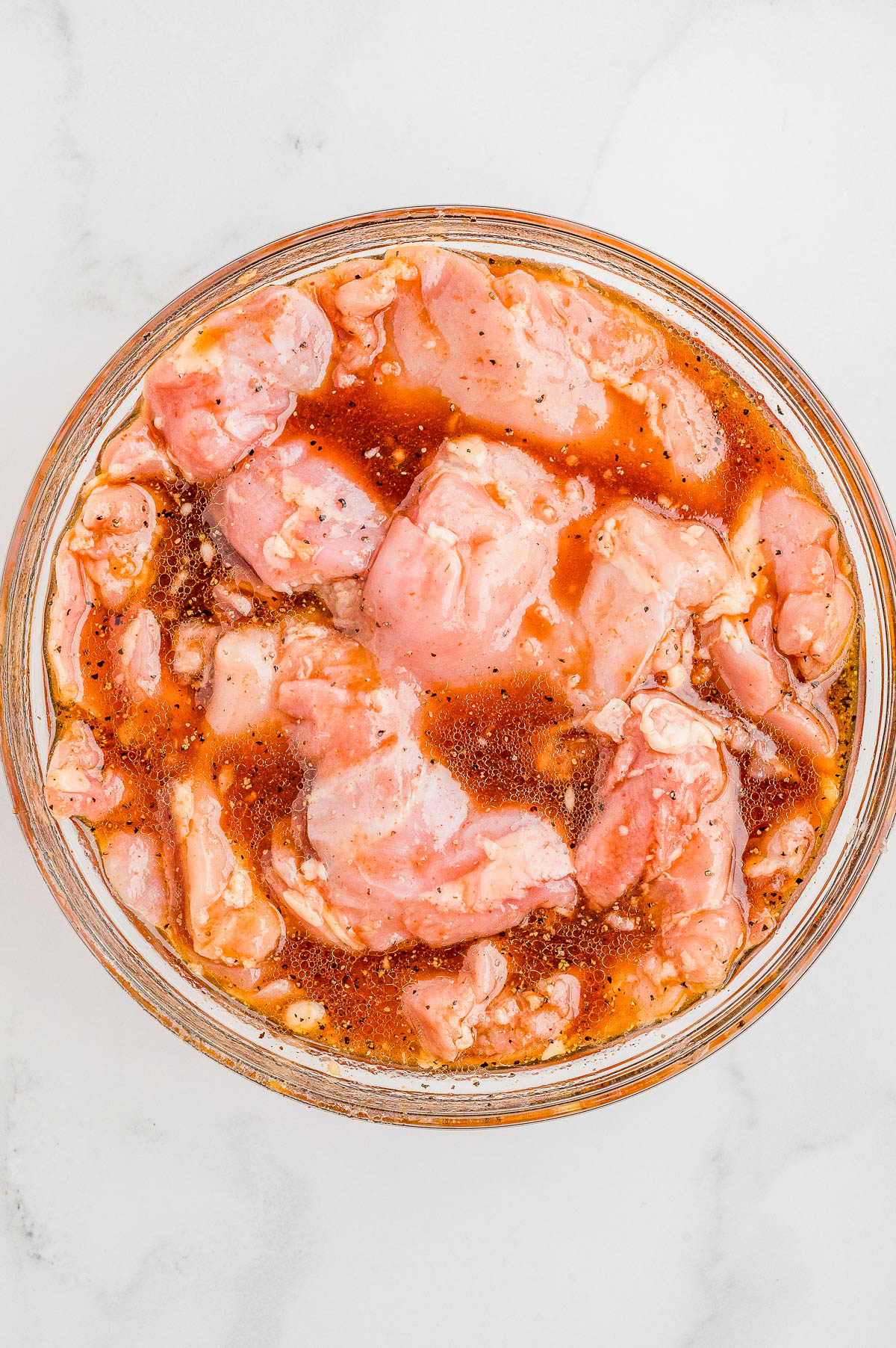 Raw chicken pieces marinating in a bowl with a reddish marinade on a white surface.