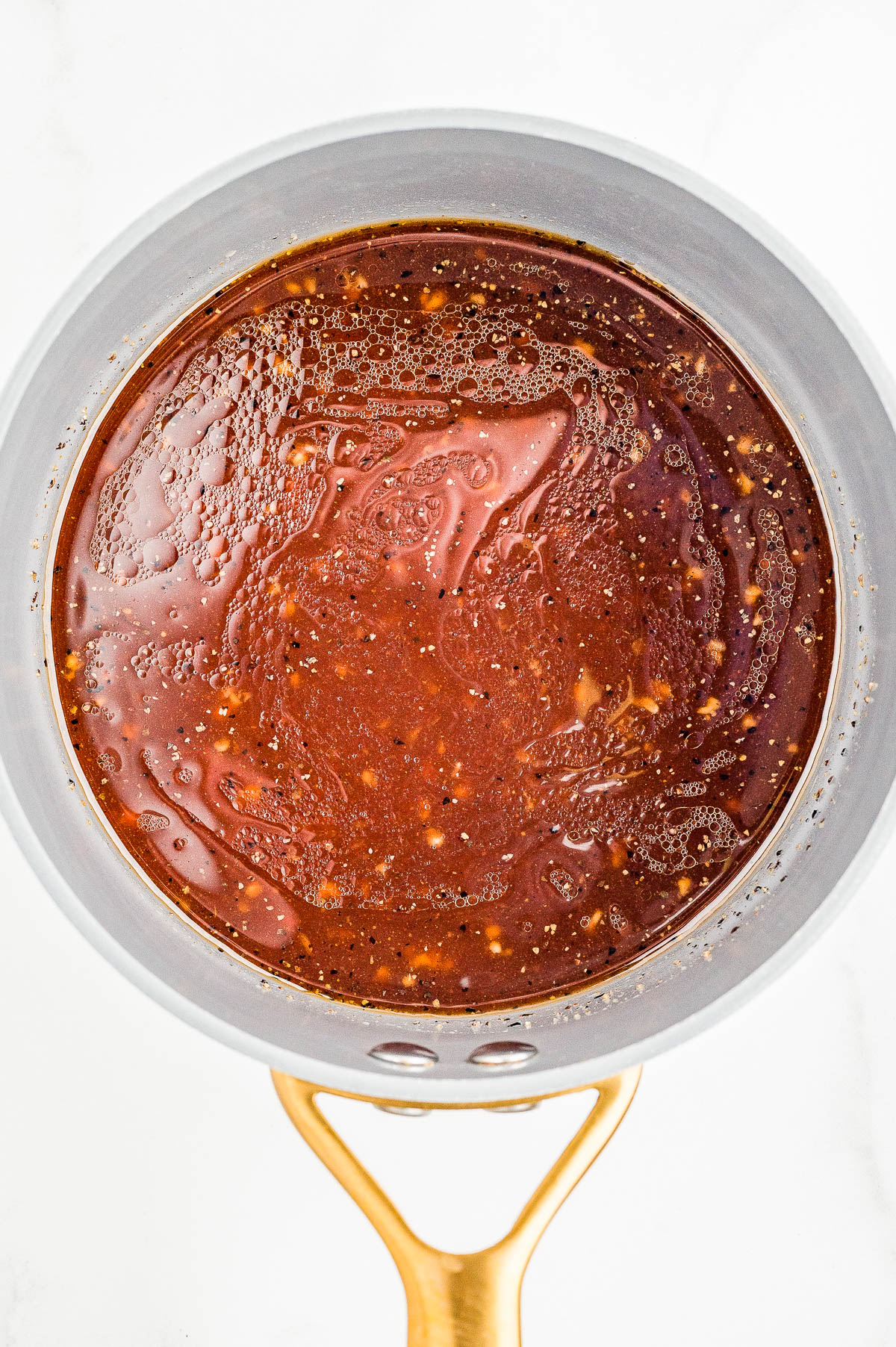 A saucepan filled with a thick, dark reddish-brown sauce containing visible seasonings and small bits of ingredients.