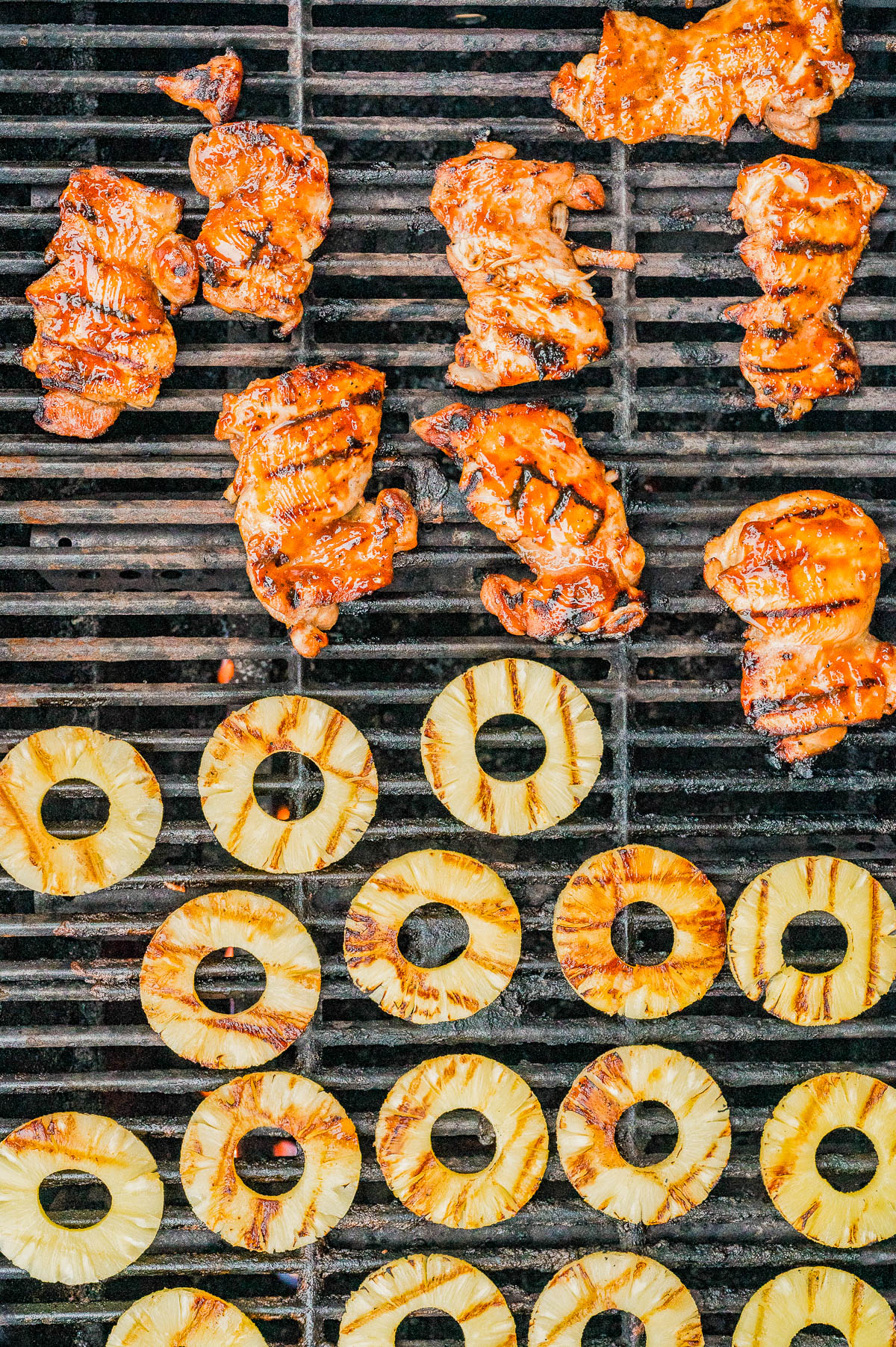 Chicken pieces and pineapple rings are being grilled on a barbecue, with the chicken on the top half and the pineapple on the lower half of the grill.