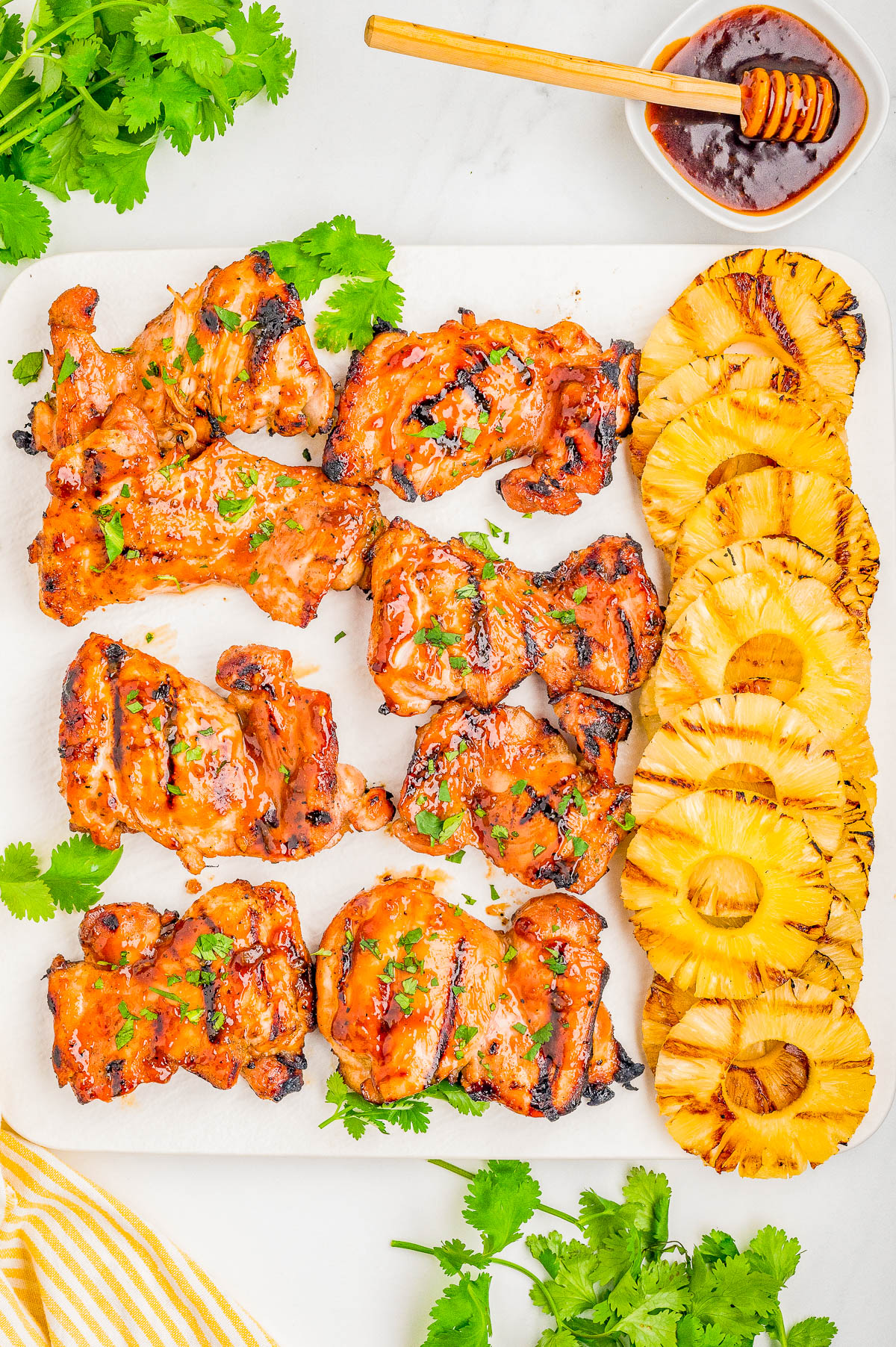 Grilled chicken thighs with glaze are arranged on a platter next to sliced grilled pineapple and fresh cilantro, with a small bowl of sauce and honey dipper nearby.