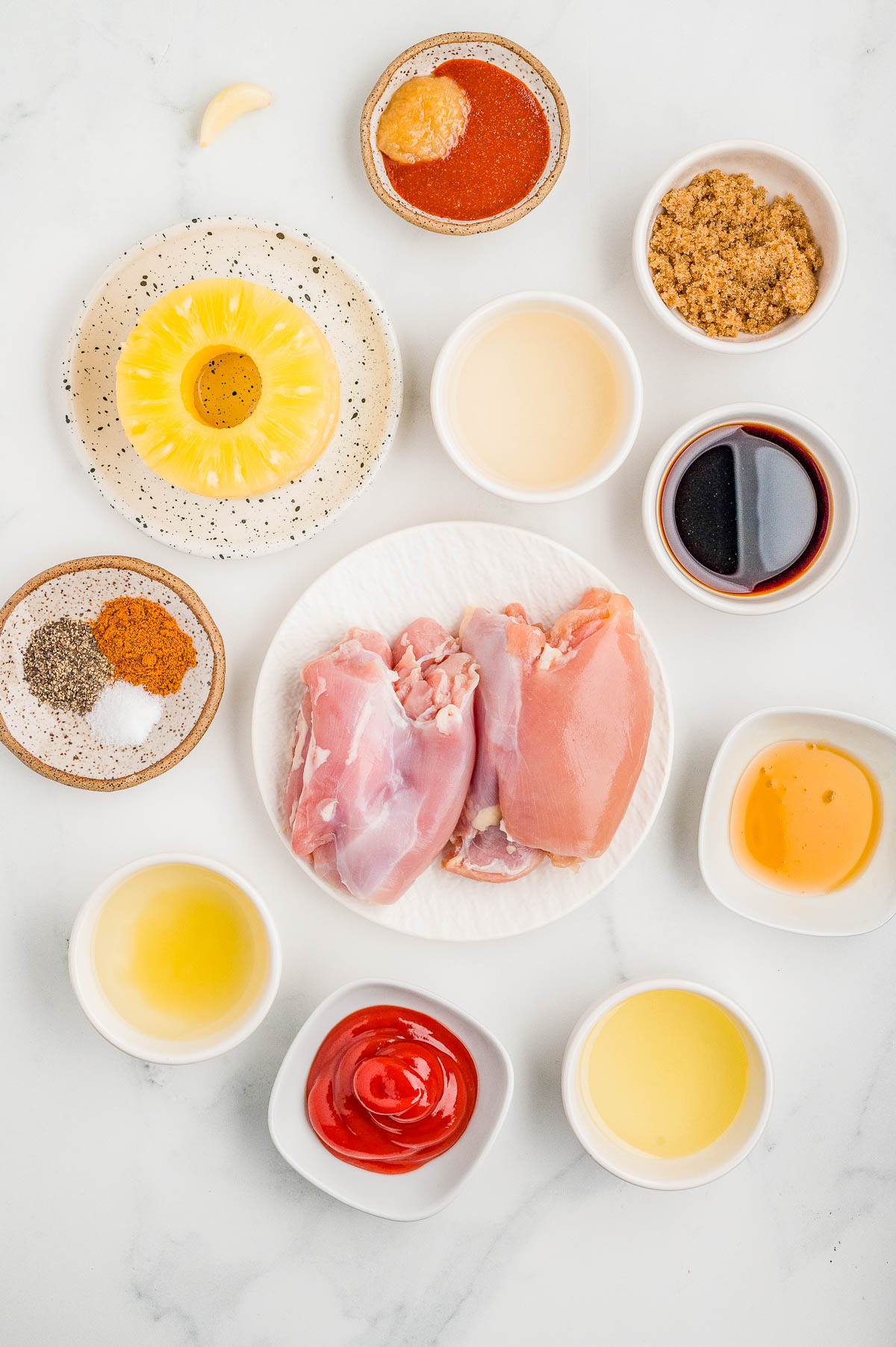 A variety of ingredients in small bowls surround raw chicken thighs on a white plate; items include pineapple, sauces, spices, and brown sugar on a white surface.