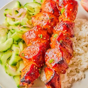 Two skewers of glazed grilled chicken served on white rice with a cucumber salad on a white plate.