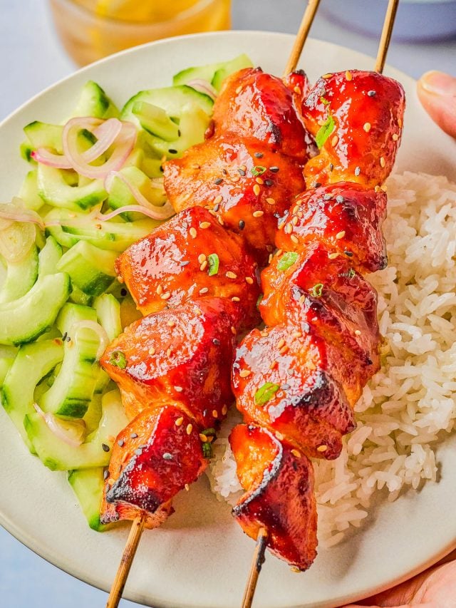 Two skewers of glazed grilled chicken served on white rice with a cucumber salad on a white plate.