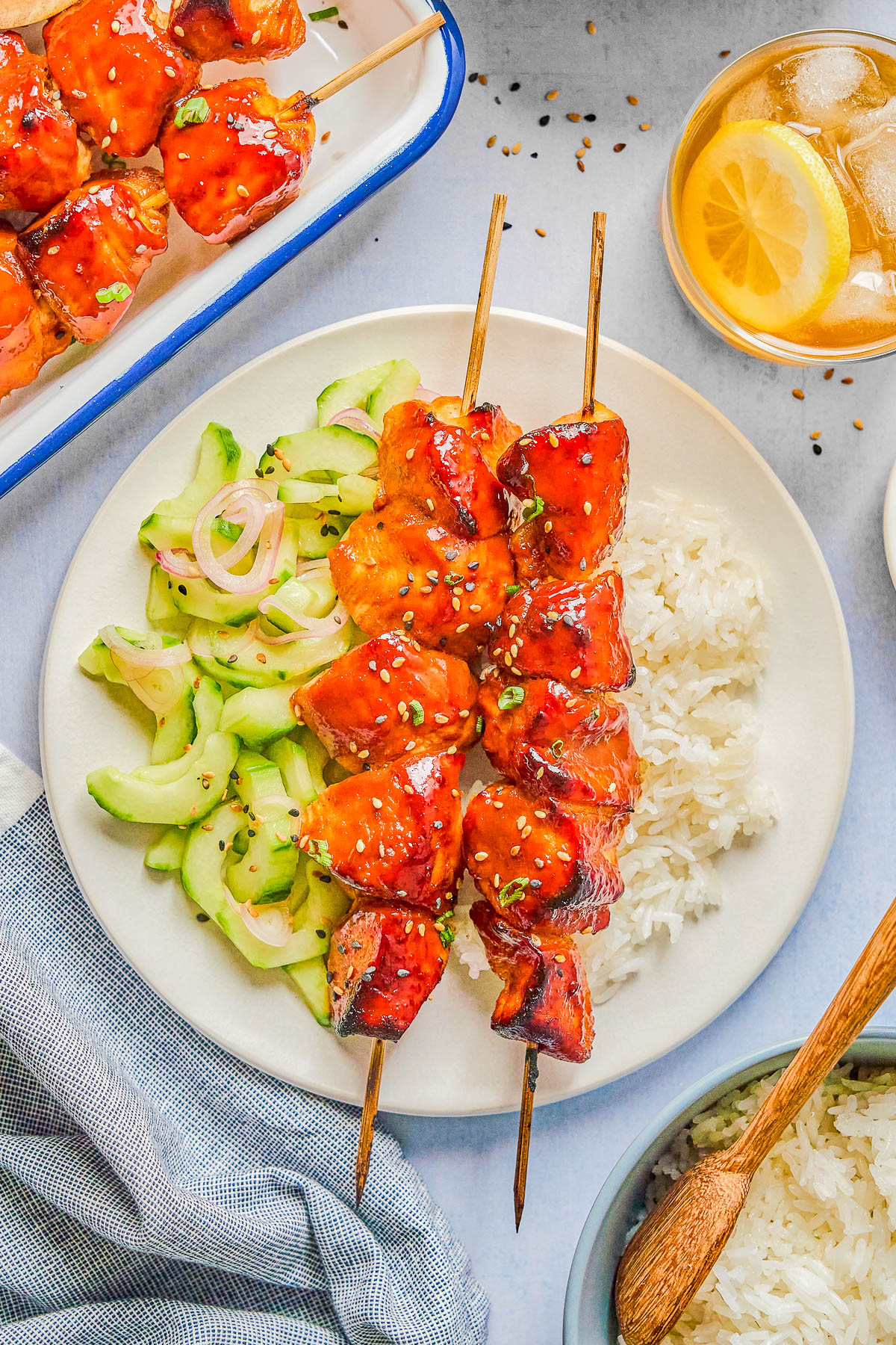 A plate with two glazed chicken skewers, white rice, and cucumber salad. Nearby are a bowl of rice, a drink with lemon, and extra chicken skewers on a tray.