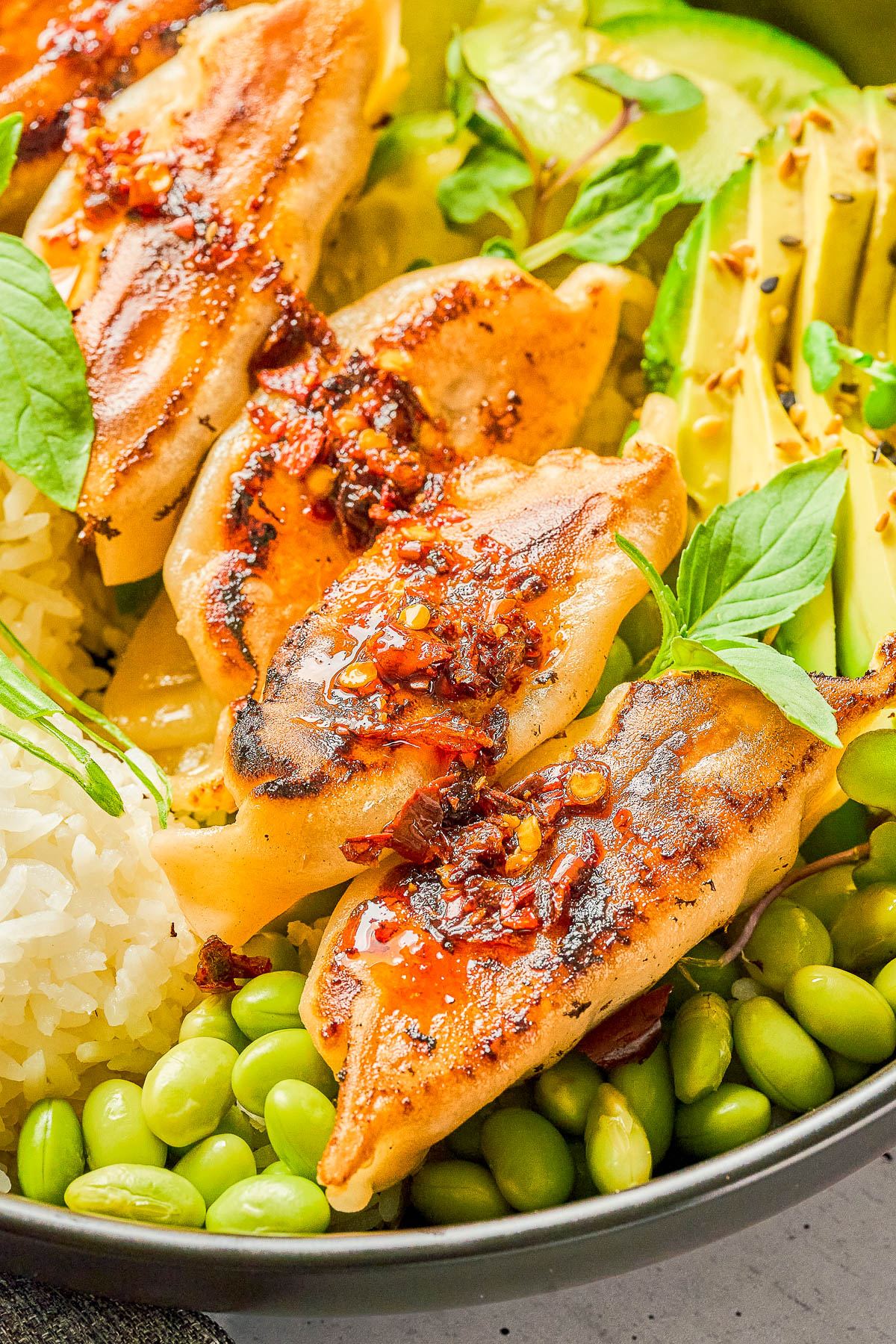 Close-up of a bowl with grilled dumplings topped with chili sauce, sliced avocado, white rice, edamame, and fresh basil leaves.