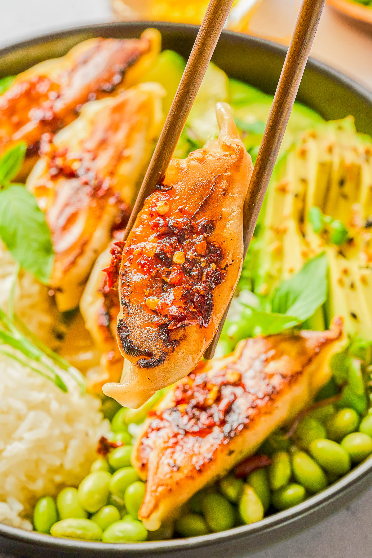 A close-up of a chopstick holding a pan-fried dumpling topped with chili sauce, with rice, edamame, sliced avocado, and more dumplings visible in the background.