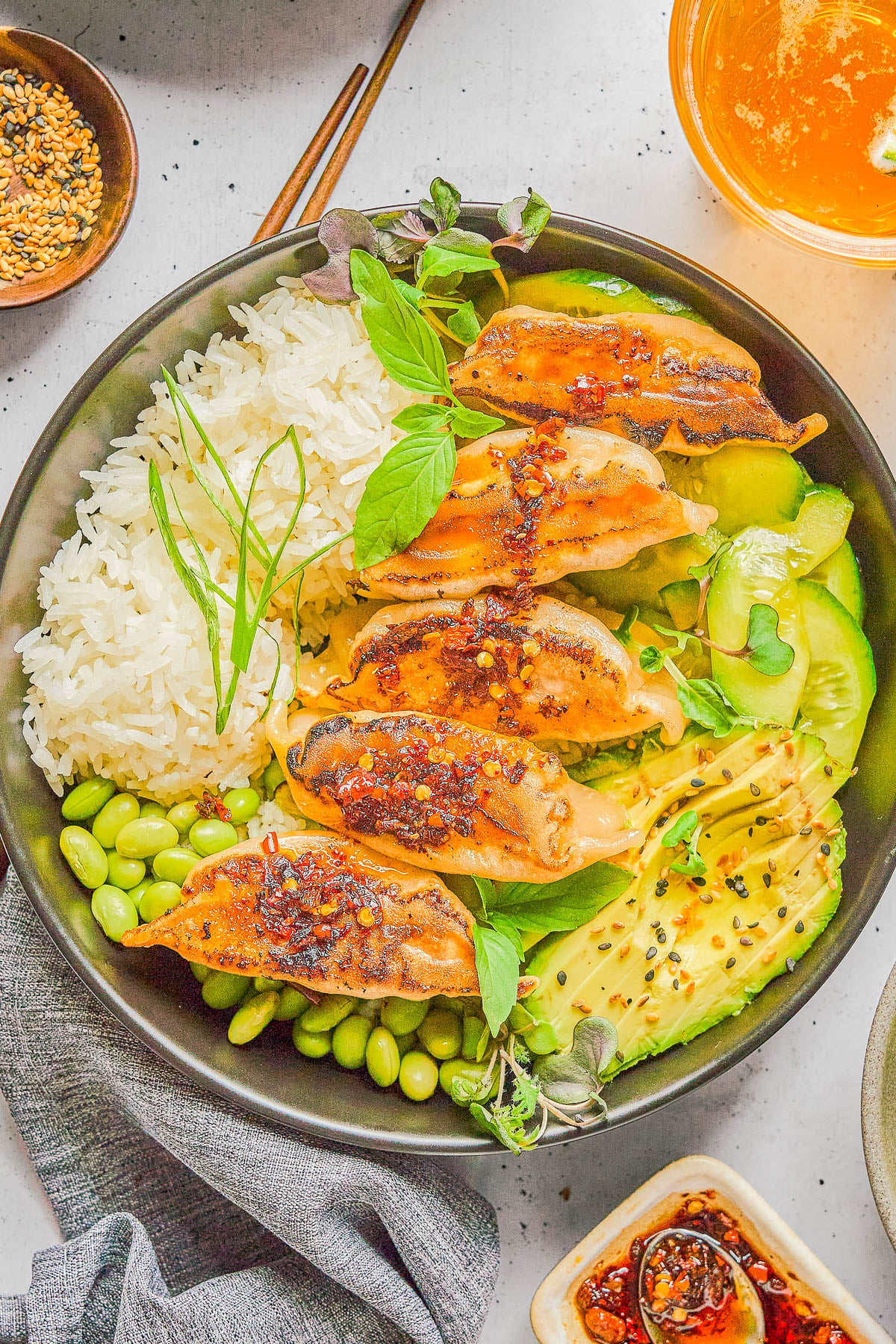 A bowl with five pan-fried dumplings, white rice, sliced avocado, edamame, cucumber, microgreens, and a drizzle of chili oil, served with a small bowl of soup and toppings on the side.