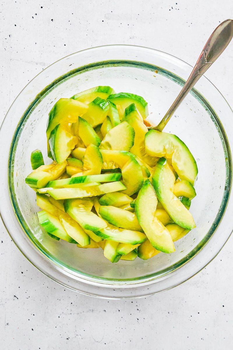 Glass bowl filled with sliced cucumbers in a yellowish dressing, with a metal spoon resting inside the bowl.