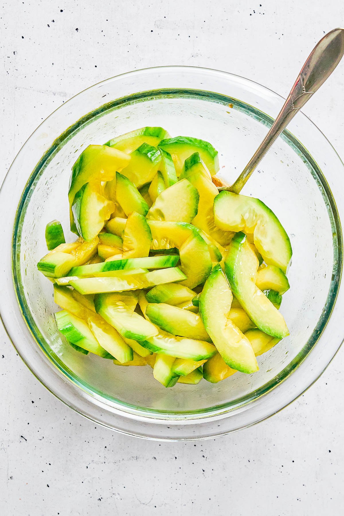 Glass bowl filled with sliced cucumbers in a yellowish dressing, with a metal spoon resting inside the bowl.