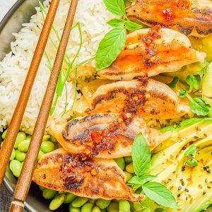 A bowl with steamed rice, five pan-fried dumplings topped with chili sauce, edamame, avocado slices, and fresh herbs, with wooden chopsticks resting on the bowl.