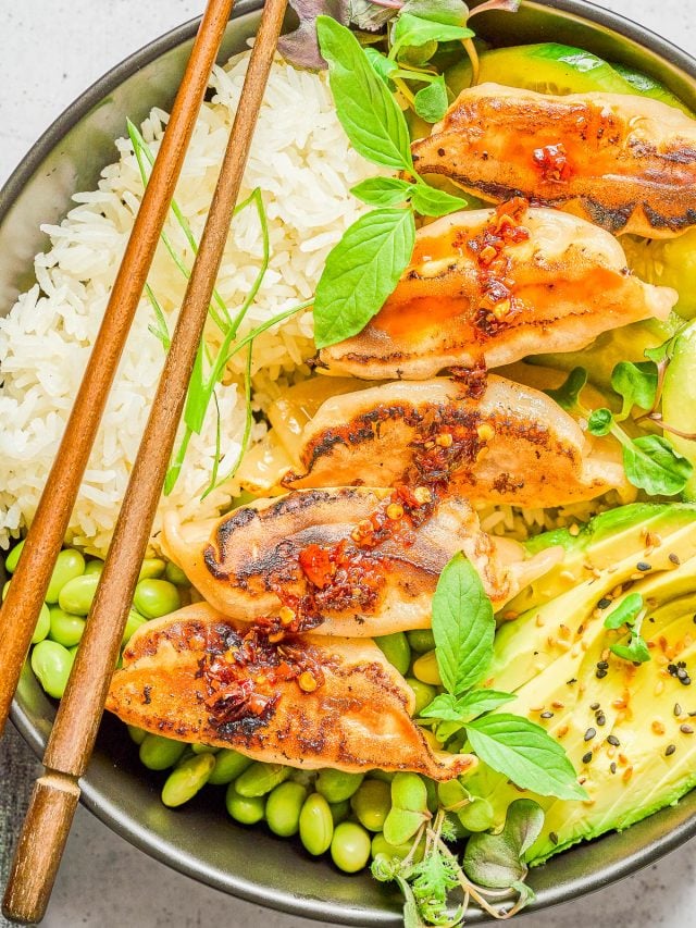 A bowl with steamed rice, five pan-fried dumplings topped with chili sauce, edamame, avocado slices, and fresh herbs, with wooden chopsticks resting on the bowl.