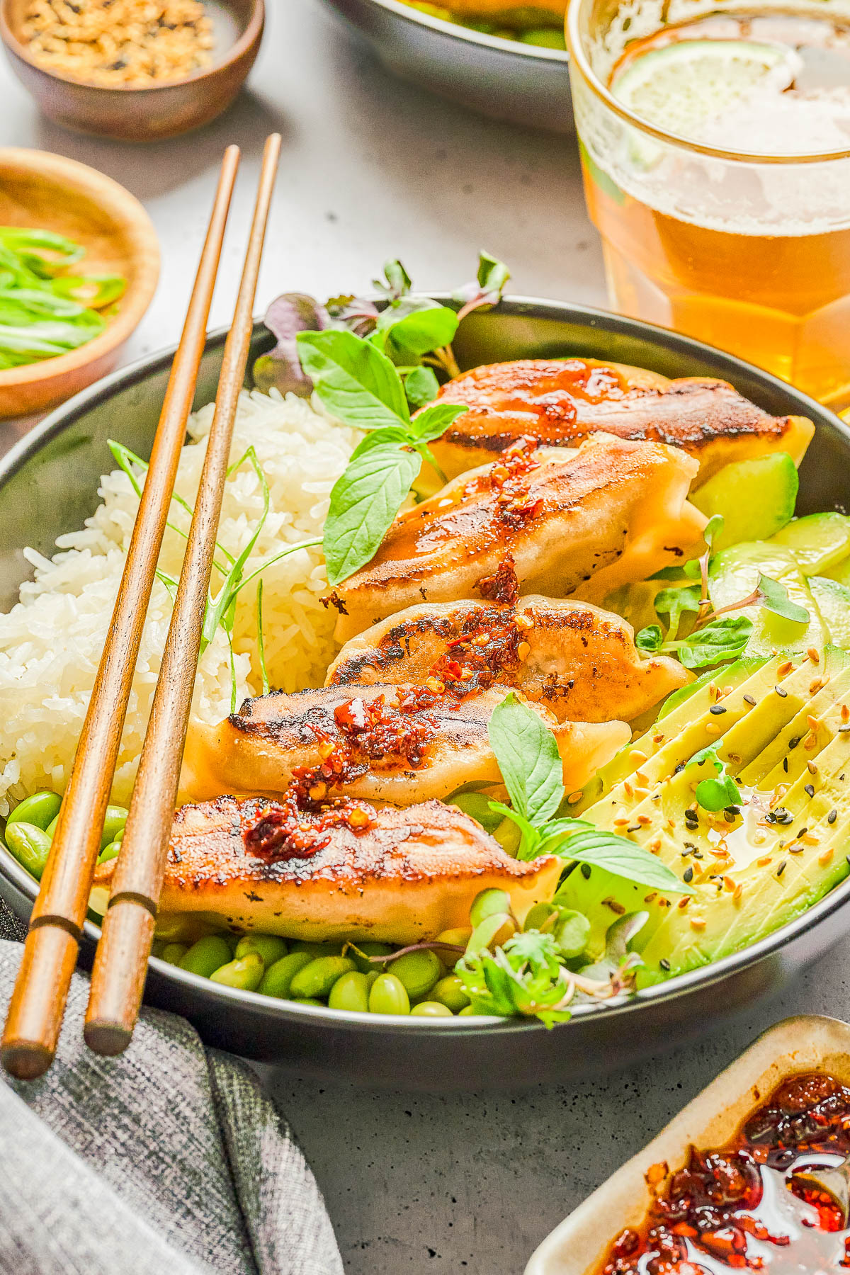 A bowl with white rice, grilled dumplings topped with chili oil, sliced avocado, edamame, fresh herbs, and wooden chopsticks.
