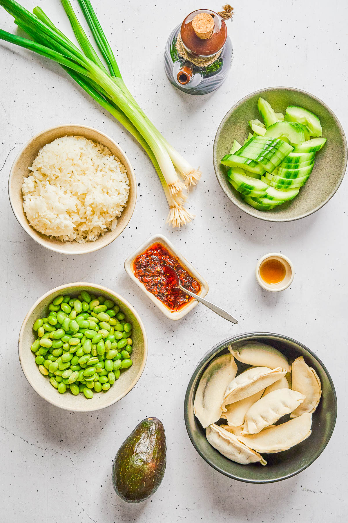 A flat lay of scallions, soy sauce, cucumber slices, rice, edamame, chili sauce, vinegar, avocado, and dumplings on a white surface.