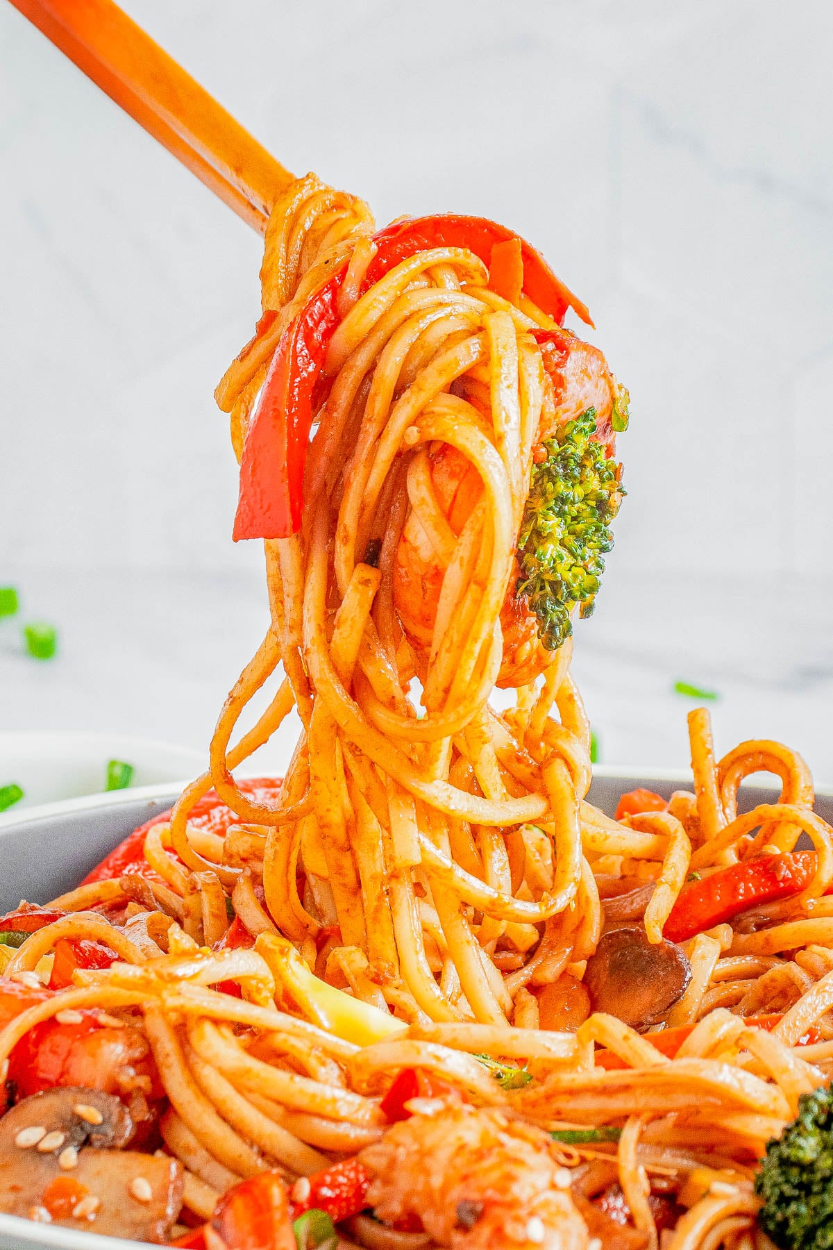 Close-up of chopsticks lifting spaghetti with vegetables, including broccoli, red bell pepper, and mushrooms, coated in a red sauce.
