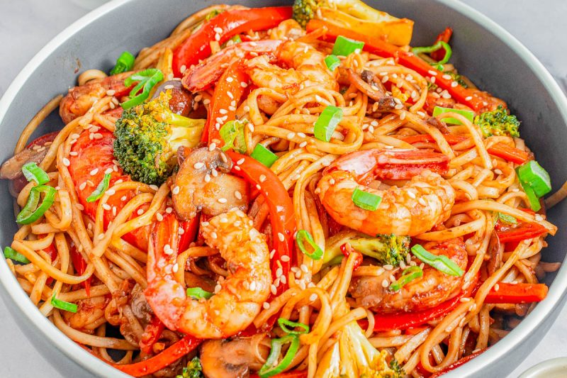 Bowl of stir-fried noodles with shrimp, red bell peppers, broccoli, mushrooms, and green onions, topped with sesame seeds.