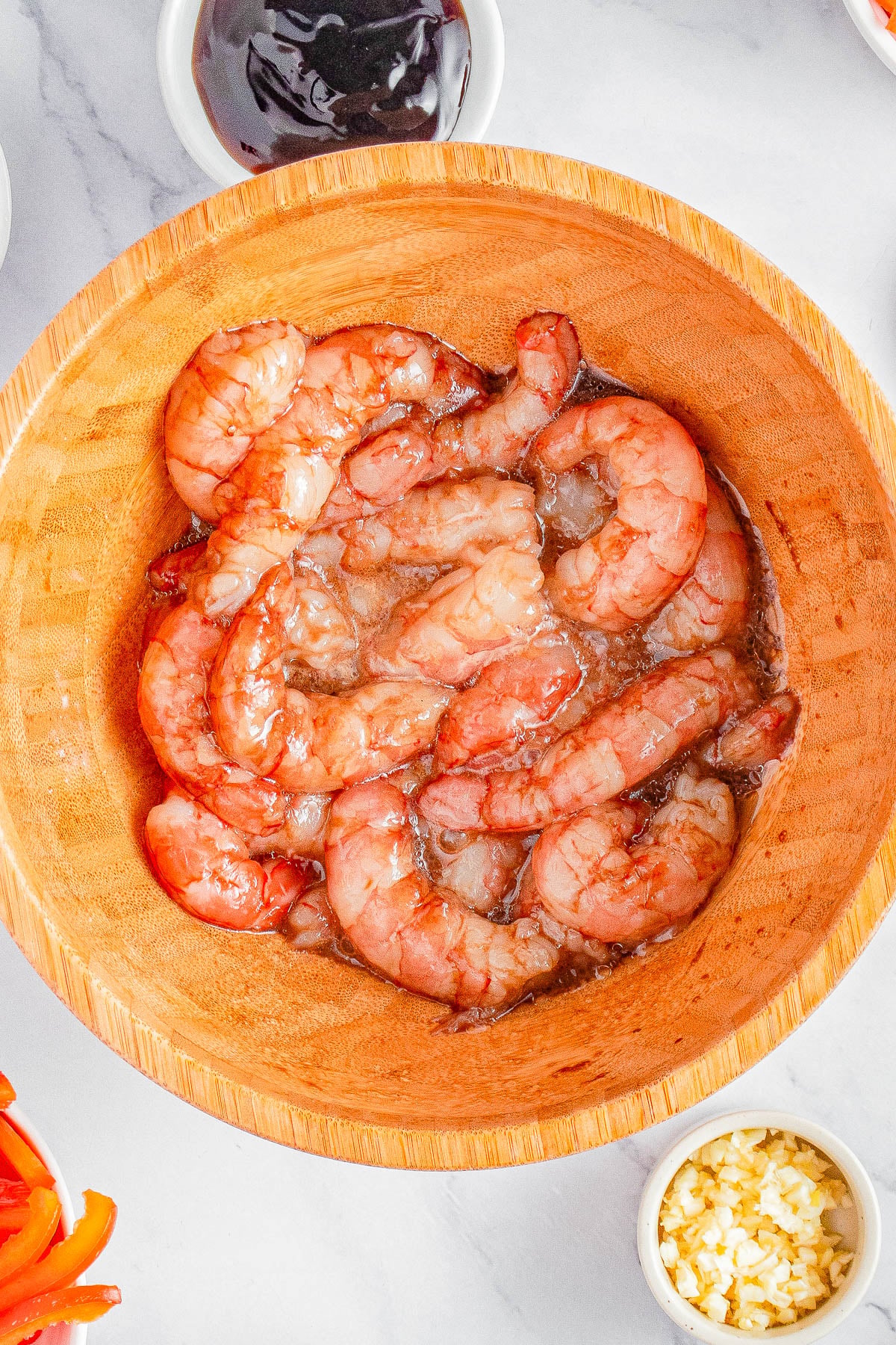 Raw shrimp marinating in a wooden bowl, surrounded by small bowls containing dark sauce, chopped garlic, and sliced vegetables.