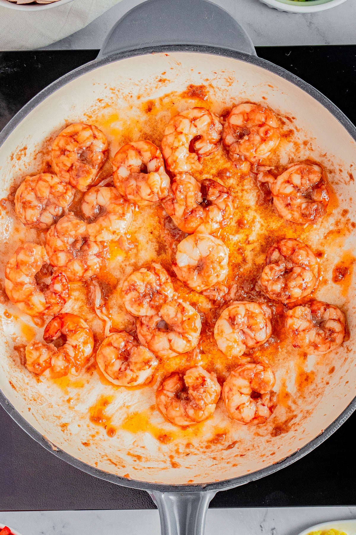 Shrimp being sautéed in a white skillet with a reddish sauce, surrounded by some oil and seasoning.