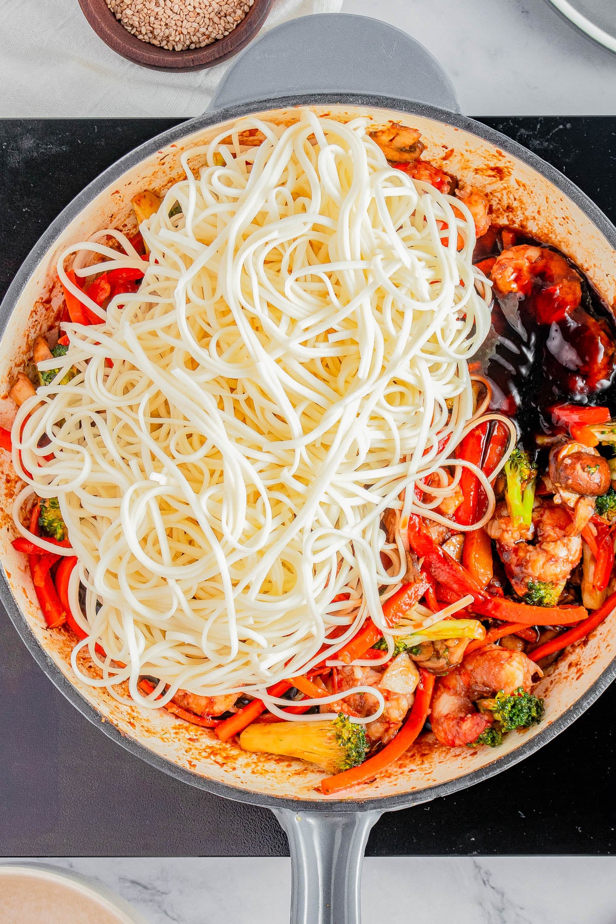 A skillet containing cooked noodles, stir-fried vegetables, pieces of chicken, and sauce, all partially mixed together on a stovetop.