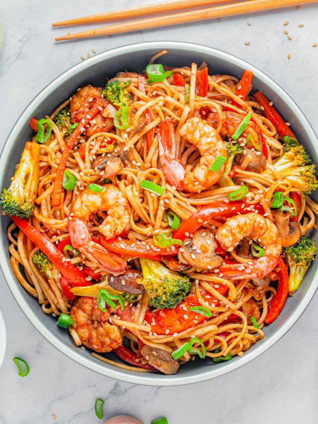 A bowl of shrimp lo mein with noodles, broccoli, red bell peppers, mushrooms, green onions, and sesame seeds, with chopsticks placed above the bowl.