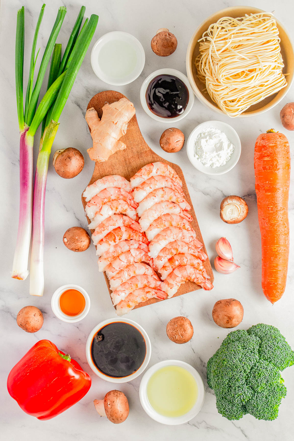 A top-down view of raw shrimp, noodles, broccoli, carrots, mushrooms, red bell pepper, green onions, ginger, sauces, cornstarch, and garlic arranged on a white surface.