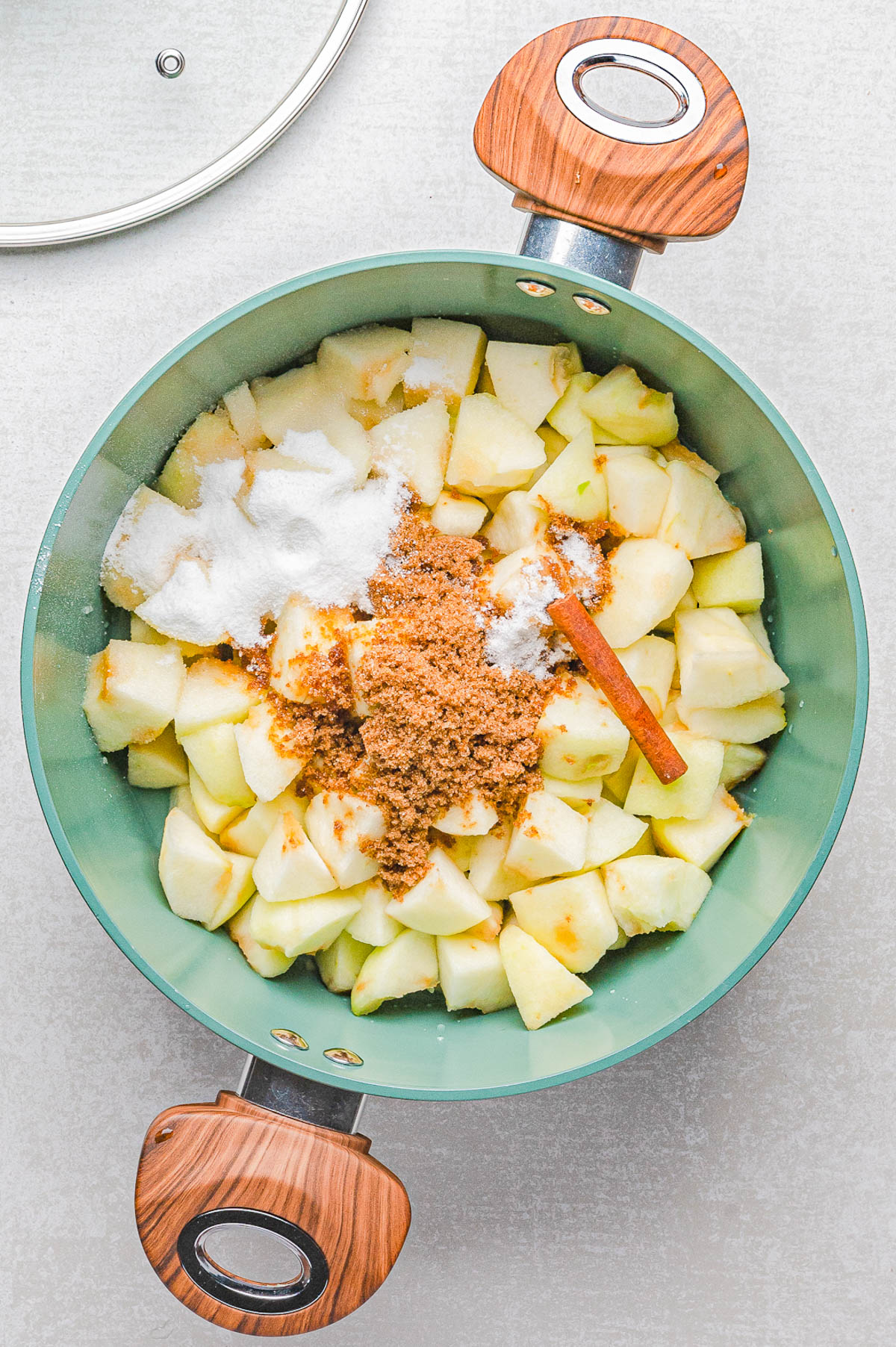 Chopped apples in a pot topped with white sugar, brown sugar, spices, and a cinnamon stick, ready for cooking.
