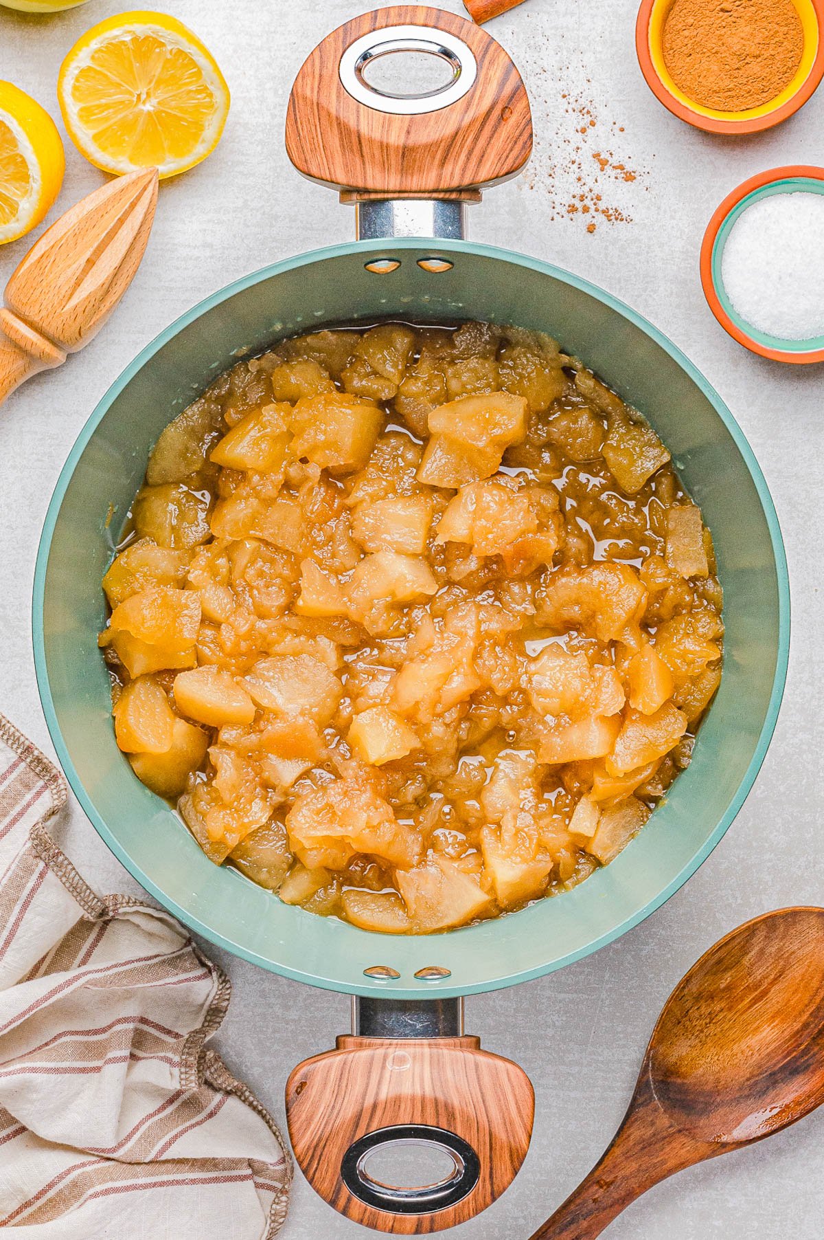 A pot filled with chunky cooked apples sits on a countertop, surrounded by a wooden spoon, a striped cloth, halved lemons, cinnamon, and sugar.