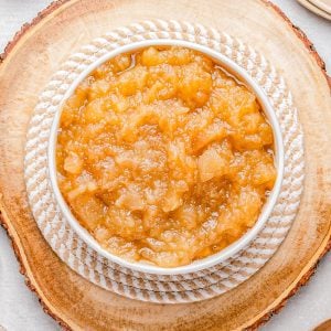 A white bowl filled with chunky applesauce sits on a wooden slab, with plates, a napkin, and cinnamon sticks visible around it.