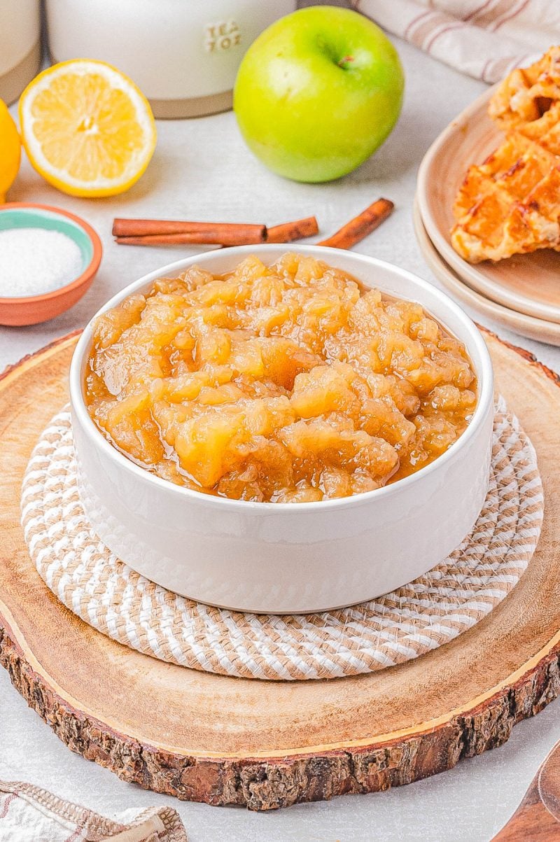A white bowl filled with chunky applesauce sits on a wooden slice, with a green apple, lemon, cinnamon sticks, and waffles in the background.