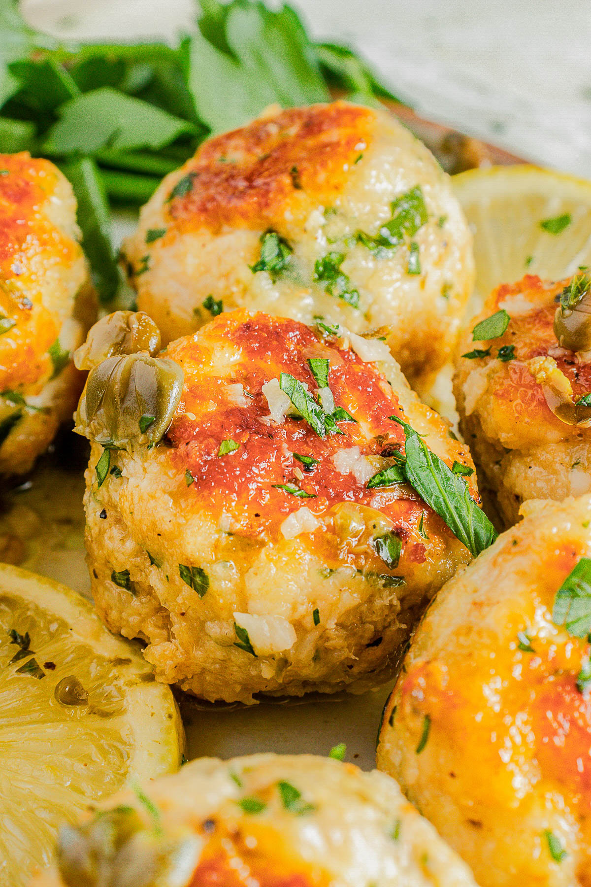 Close-up of baked chicken meatballs garnished with chopped herbs and capers, with lemon slices and parsley in the background.