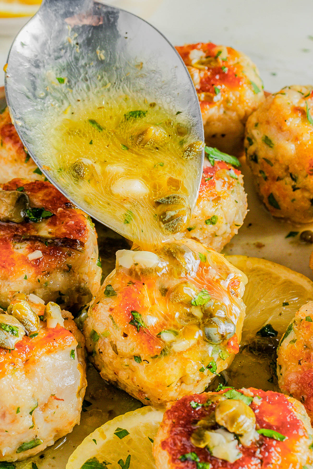 A spoon drizzles sauce over golden-brown meatballs garnished with herbs and capers, with lemon slices nearby.