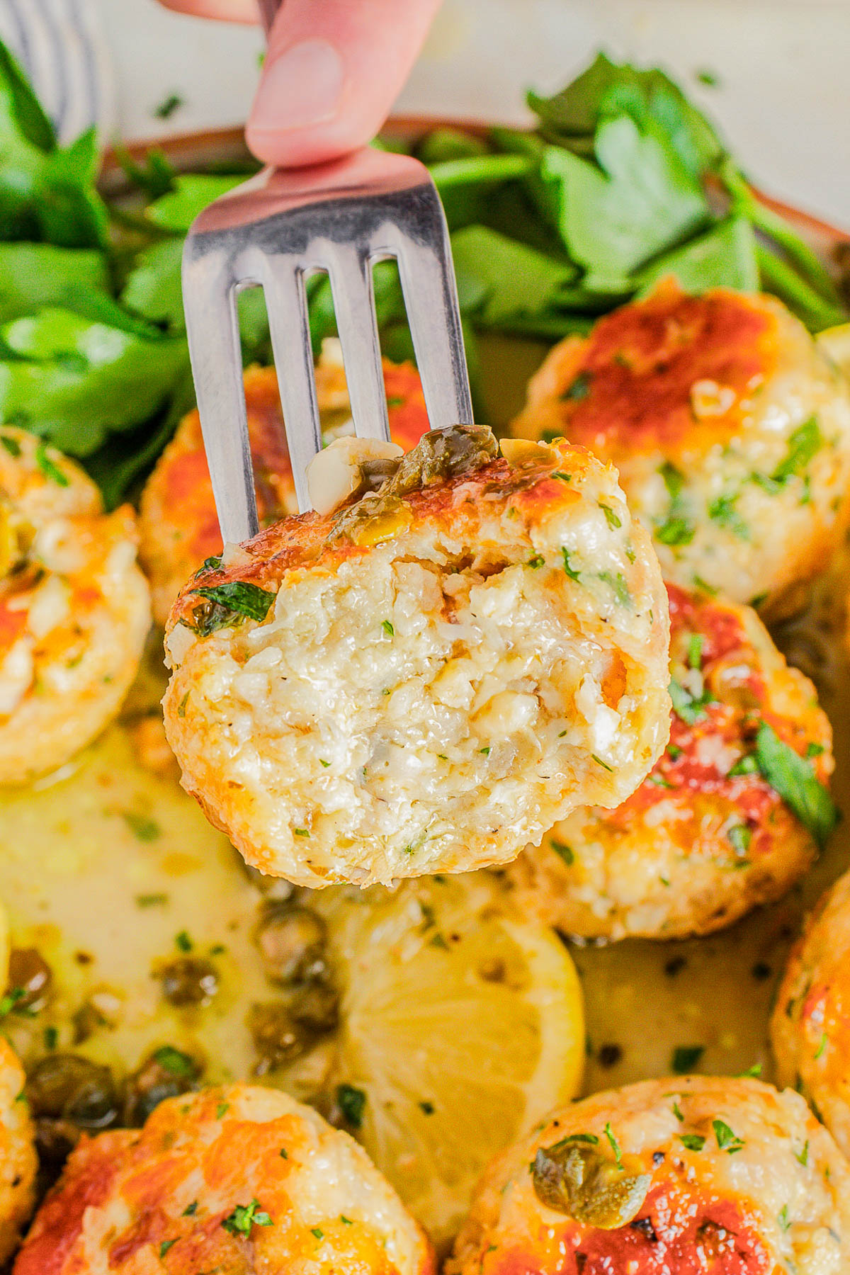 A metal spatula holding a baked fish cake garnished with herbs, with more fish cakes, lemon slices, capers, and parsley in the background.