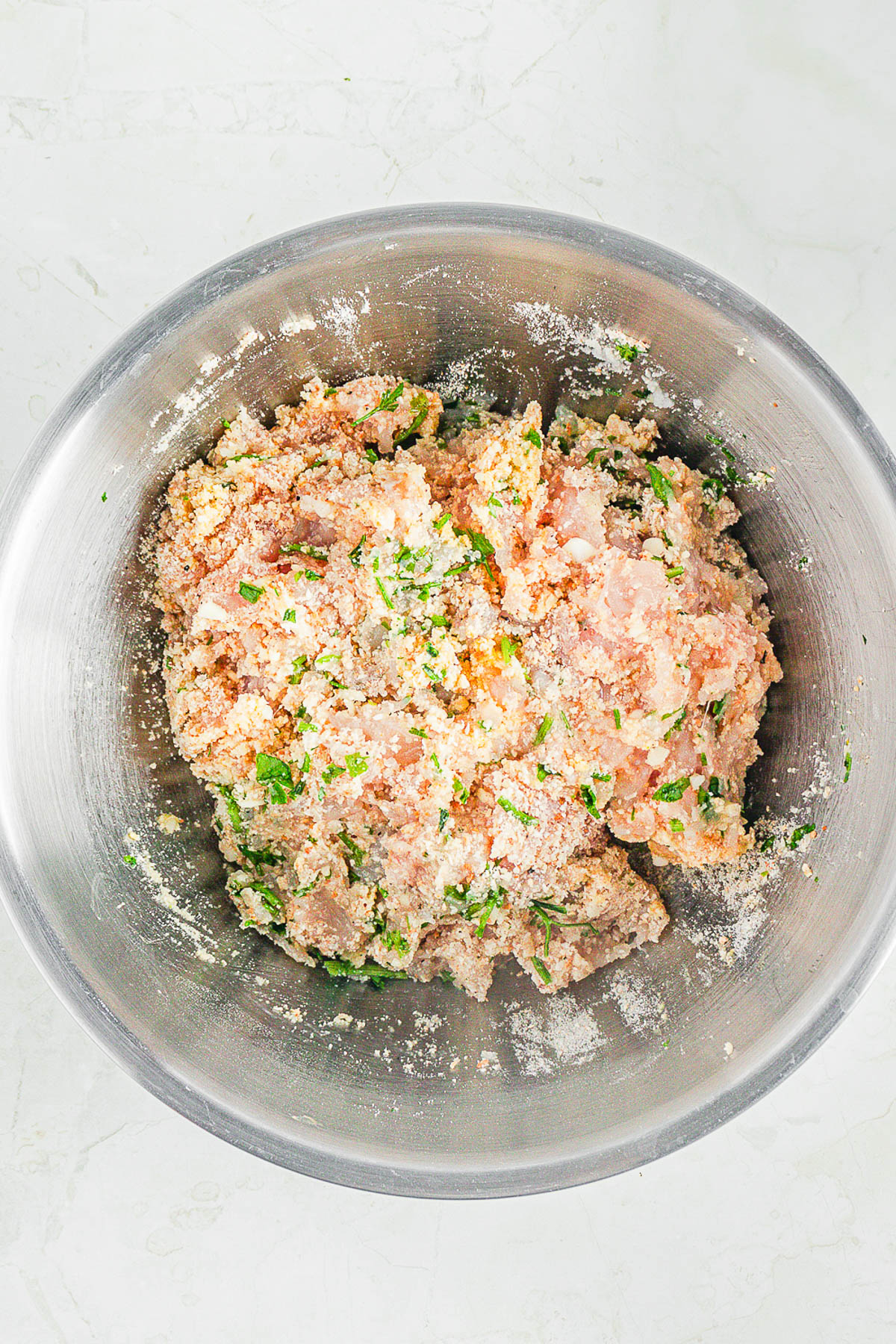 A stainless steel bowl containing a mixture of raw ground meat, herbs, and breadcrumbs on a light countertop.
