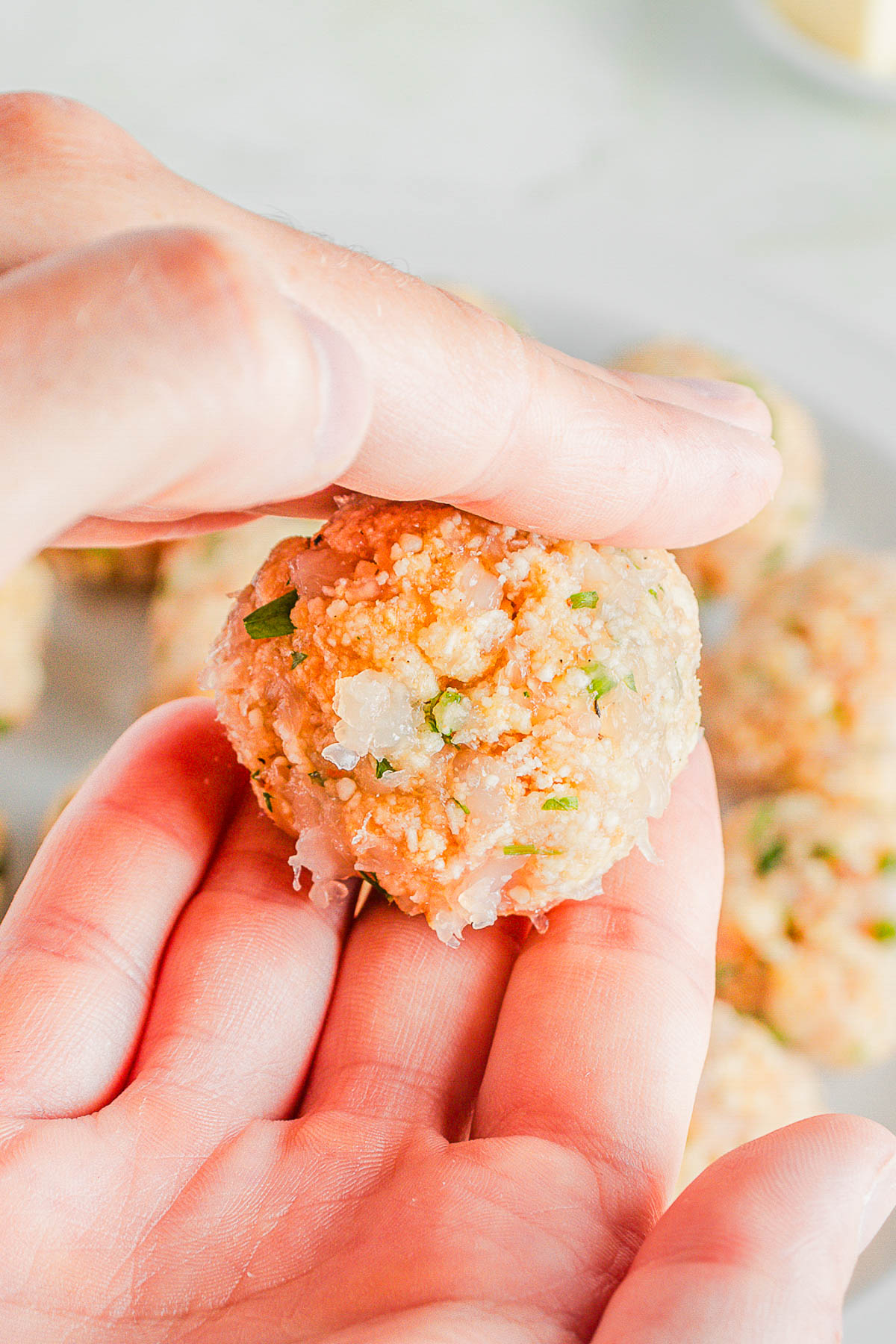 A hand holds a raw meatball mixture with visible chopped herbs and onions, while more uncooked meatballs are in the background.